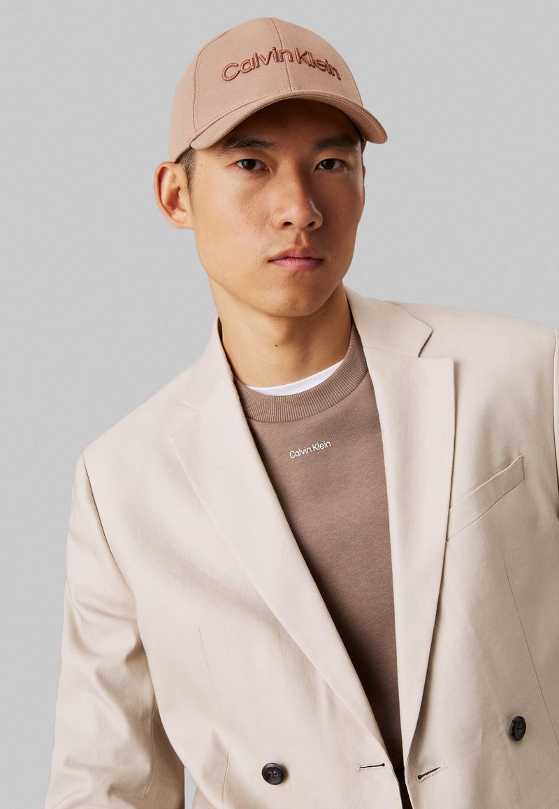 Blazer beige de doble botonadura sobre un suéter marrón de Calvin Klein, combinado con una gorra color tostado con un logotipo bordado. Texturas suaves y líneas limpias.