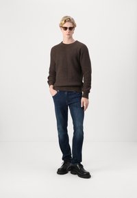 Bruine geribde gebreide trui, blauwe denim jeans en zwarte veterschoenen. Het model draagt een zonnebril en staat met de handen in de zakken.