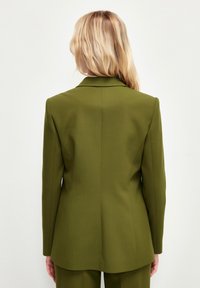 Olivgrön skräddarsydd kavaj med skarpa slag, strukturerade axlar och en slät yta, med en enda sprund bak.