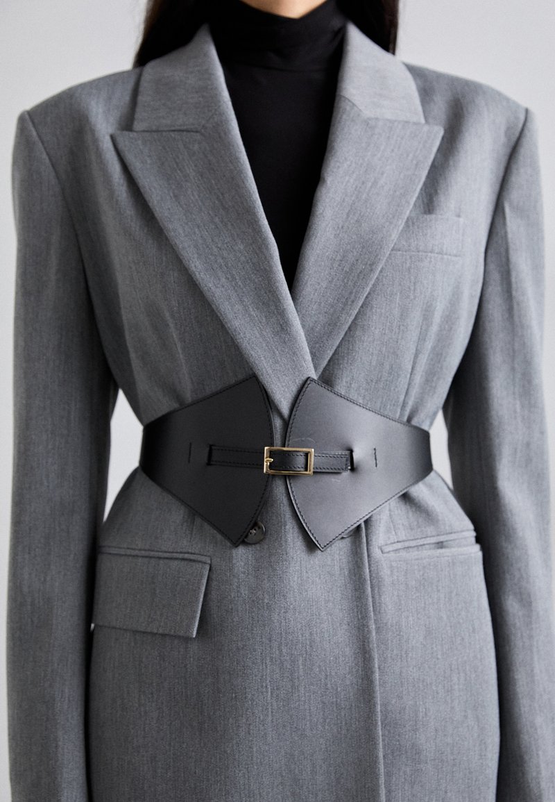 Blazer gris a medida con solapas en pico y dos bolsillos frontales, ceñido con un cinturón de cuero negro con hebilla dorada.