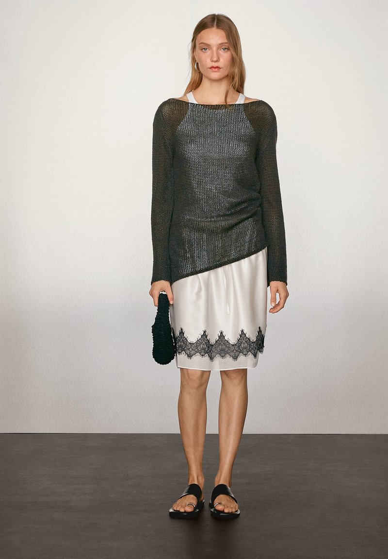 Schwarz metallisch gestricktes Oberteil mit langen Ärmeln und asymmetrischem Schnitt; cremefarbene Satin-Rock mit schwarzem Spitzenbesatz; schwarze flache Sandalen und eine strukturierte Clutch.