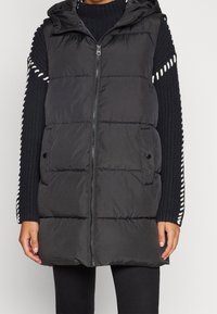 Czarna pikowana kamizelka puchowa z kapturem, z przeszyciami, z zamkiem błyskawicznym z przodu i dwoma kieszeniami bocznymi. Noszona na teksturowanym czarnym swetrze.