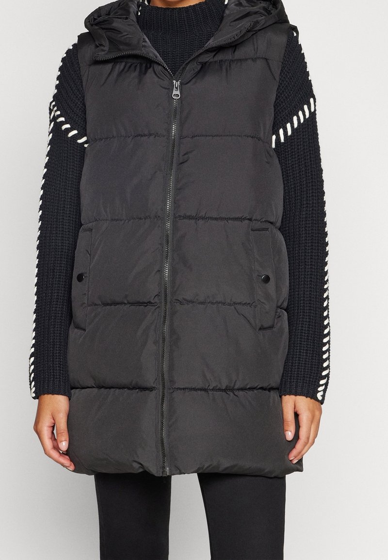 Czarna pikowana kamizelka puchowa z kapturem, z przeszyciami, z zamkiem błyskawicznym z przodu i dwoma kieszeniami bocznymi. Noszona na teksturowanym czarnym swetrze.