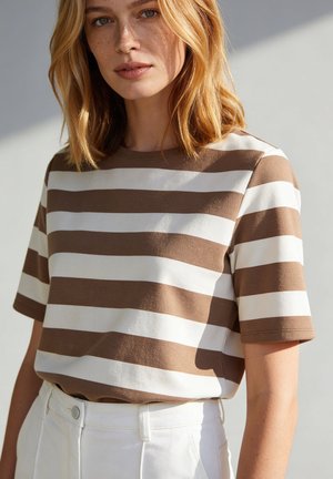 Vrouw met schouderlang blond haar die een bruin-wit horizontaal gestreept shirt met korte mouwen en een witte broek draagt tegen een effen achtergrond.