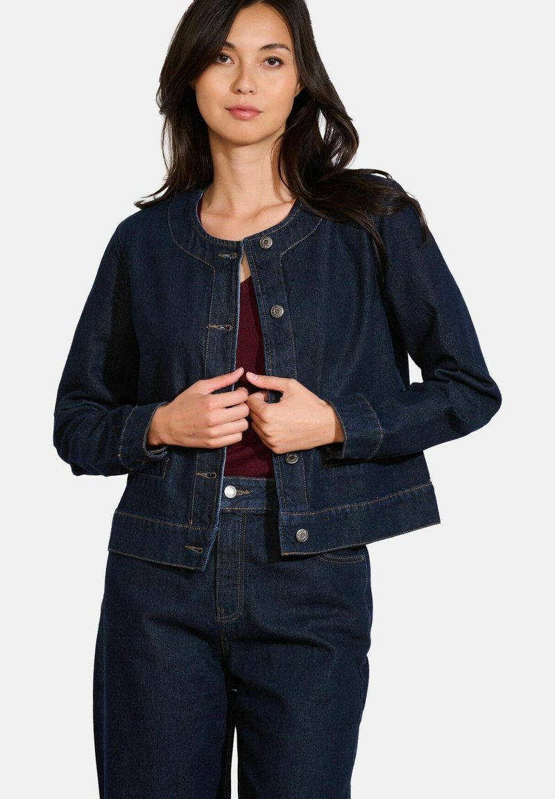 Veste en denim foncé avec un col rond, fermeture à boutons-pression, deux poches avant et des coutures contrastantes. Portée sur un haut bordeaux.