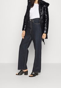 Czarna kurtka puchowa, biała koszulka, ciemnoniebieskie dżinsy o wysokim stanie i szerokich nogawkach, oraz czarne klapki na słupku z otwartym palcem. Widoczne kontrastowe przeszycia.