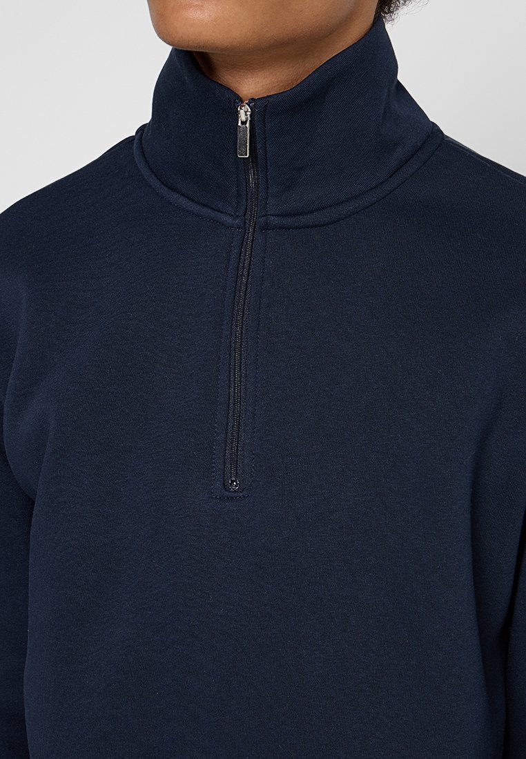 Pull bleu marine avec un col montant et une fermeture éclair à mi-hauteur. Fabriqué en tissu doux avec une texture lisse et un minimum de détails de couture.