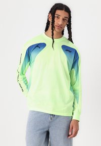 Langermet, neon grønn netting skjorte med blå gradient detaljer og en svart logo på brystet og ermet, med en løs passform.