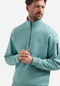 Lichtblauwe sweatshirts met rits en halsmade van zachte stof. Heeft een klein logo-patch en een zijrits zak op de mouw.