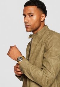 Veste en simili cuir olive avec un col montant, dotée d'une fermeture éclair à l'avant et de boutonnières ; montre noire et or au poignet.