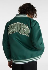 Vihreä pommikuri, jossa on suuri valkoinen brodeerattu "VANS"-logo selässä, raidalliset ribbikujat ja sileä, kiiltävä pinta.