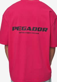 Rózsaszín pamut póló, fekete grafikus nyomattal a hátsó részén, amelyen a "PEGADOR" és a "CERTIFIED AUTHENTIC STREETWEAR" felirat szerepel. Rövidujjú, laza szabású.