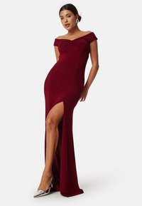Burgundy aftonklänning med off-shoulder-design, frontslits och figurnära silhuett i ett glittrande material. Silverfärgade spetsiga klackar framhäver looken.