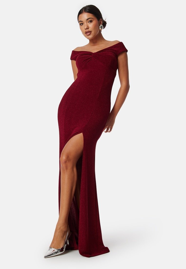 Burgundy aftonklänning med off-shoulder-design, frontslits och figurnära silhuett i ett glittrande material. Silverfärgade spetsiga klackar framhäver looken.