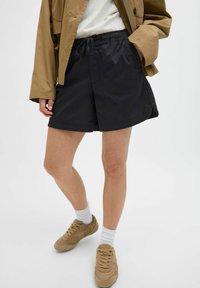 Pantalones cortos negros con cintura elástica y cordón, que cuentan con bolsillos laterales. Combinados con una chaqueta beige y zapatillas de color marrón claro.