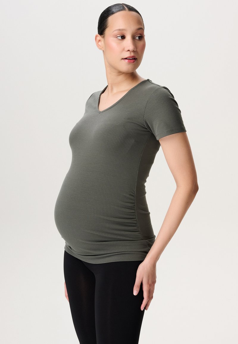 Graviditetsbluse i ribbet olivengrønt stof, med v-hals, korte ærmer og samlede detaljer i siderne, båret med sorte leggings.