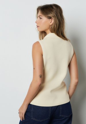 Mujer con cabello a la altura de los hombros, vestida con una camiseta sin mangas beige y jeans azul oscuro, mostrando tatuajes en su brazo izquierdo, sobre un fondo liso.