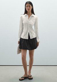 Camicia bianca a maniche lunghe con bottoni e finitura testurizzata, abbinata a una gonna nera con coulisse e infradito nere. Borsa intrecciata in mano.