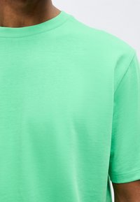 Κοντινό ενός ατόμου που φορά ένα απλό, μέντας πράσινο T-shirt με κοντά μανίκια και στρογγυλή λαιμόκοψη σε ανοιχτό φόντο.