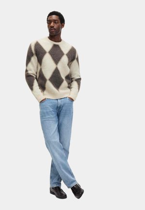 Hombre vestido con un suéter argyle en crema y marrón, jeans azul claro y zapatos negros, de pie con las manos en los bolsillos y las piernas cruzadas.