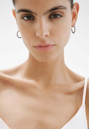 Aros de plata en forma de aro con detalles en espiral, usados por un modelo con piel suave, maquillaje mínimo y una blusa blanca sencilla.