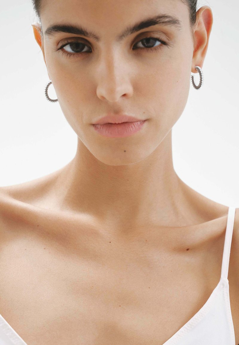Aros de plata en forma de aro con detalles en espiral, usados por un modelo con piel suave, maquillaje mínimo y una blusa blanca sencilla.