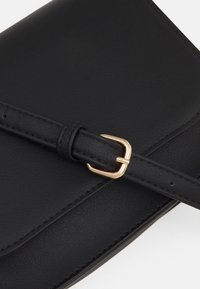 Borsa a mano in pelle nera con una texture liscia, caratterizzata da una fibbia e dettagli della tracolla in tonalità oro. Design rettangolare semplice con bordi cuciti.