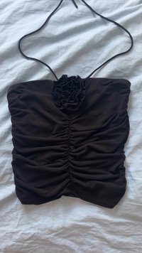 Haut noir froncé avec un accent floral, en tissu doux. Présente des bretelles fines et une forme ajustée, posée à plat sur un fond clair.