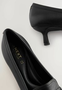 Scarpe nere in pelle con tacco alto, punta tondeggiante e tacco sottile. Presentano cuciture discrete e il marchio "NEXT MOTIONFLEX" sulla soletta.