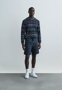 Tamsiai mėlynas megztinis su raštuotu mezgimu, derinamas su tamsiais šortais. Modelis dėvi baltus sportbačius ir kojines. Minimalistinis fonas.