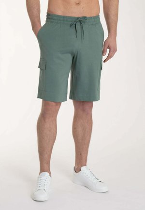 Uomo che indossa pantaloncini cargo verdi con coulisse in vita e sneakers bianche, in piedi davanti a uno sfondo semplice.