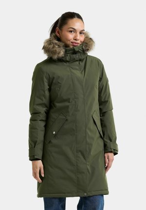 Femme portant un manteau d'hiver vert avec une capuche bordée de fourrure, debout les mains détendues, sur un fond clair uni.