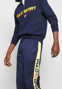 Marinefarbener Reißverschluss-Sweater mit "POLO SPORT" in leuchtendem Gelb und Rot, kombiniert mit marineblauen Jogginghosen mit einem gelben Seitenstreifen und Logo.