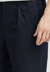 Sötétkék nadrág, texturált anyagból, plisszírozott dizájnnal és oldalsó zsebekkel. Egy kéz finoman nyugszik az anyagon.