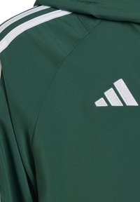 Grüne Sportjacke mit weißen Akzentstreifen, aus strukturiertem Stoff und einem gestickten weißen Logo auf der oberen linken Seite.