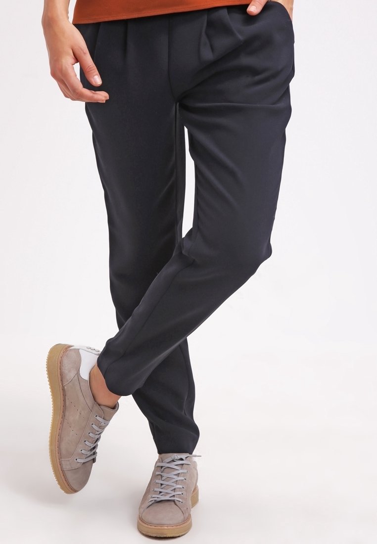 Personne portant un pantalon noir et des baskets marron clair, debout avec les jambes croisées et les mains près des poches.