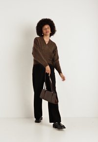 Brązowy sweter z dzianiny, czarne spodnie o szerokich nogawkach i błyszcząca brązowa torebka. Modelka ma na sobie czarne buty i stoi na tle białym.