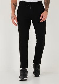 Schwarze Jogginghosen mit tailliertem Design, elastischem Bund, Kordelzugverschluss und Seitentaschen; kombiniert mit schwarzen Sneakers.