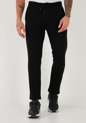 Schwarze Jogginghosen mit tailliertem Design, elastischem Bund, Kordelzugverschluss und Seitentaschen; kombiniert mit schwarzen Sneakers.