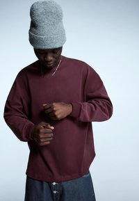 Bordowa bluza o luźnym kroju, z okrągłym dekoltem i długimi rękawami. Połączona z szarą dzianinową czapką i dżinsowymi spodniami. Subtelna tekstura.