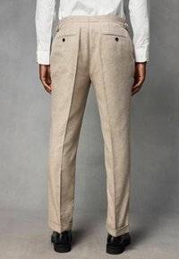 Pantalon beige sur mesure en tissu texturé, avec un devant plat, deux poches arrière et un ourlet retroussé. Chaussures habillées noires visibles.