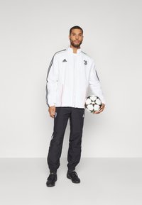 Chaqueta blanca con detalles en negro y rojo, logo en el pecho. Pantalones negros con rayas blancas y logo. Sosteniendo un balón de fútbol. Calzado negro.