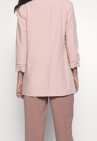 Osoba v světle růžovém blejzru s ohrnutými rukávy a odpovídajícími mauve kalhotami, zobrazená zezadu na bílém pozadí.