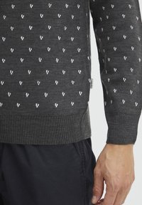 Pull en gris foncé avec un motif répétitif en petites touches blanches, des poignets et un ourlet côtelés, et une étiquette sur le côté. Texture douce.