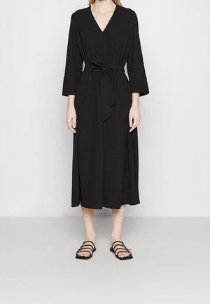 Femme portant une longue robe noire à col en V avec des manches trois-quarts et une ceinture nouée à la taille, associée à des sandales plates noires à brides.