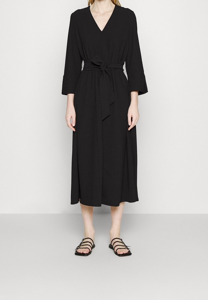 Femme portant une longue robe noire à col en V avec des manches trois-quarts et une ceinture nouée à la taille, associée à des sandales plates noires à brides.