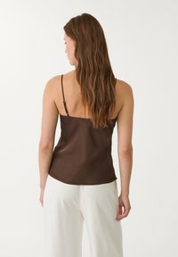 Camisole en satin marron avec des fines bretelles, présentant une texture lisse et un ourlet légèrement arrondi, portée avec un pantalon blanc.
