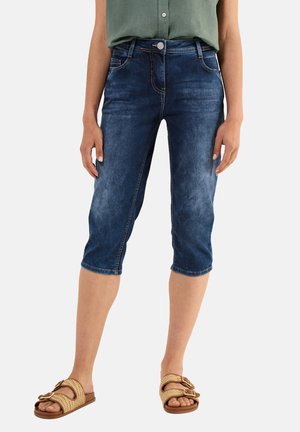 Pantalones capri de denim azul oscuro con una cintura de media altura, ligero desgaste, cinco bolsillos y acentos de costura; combinados con sandalias planas en color neutro.