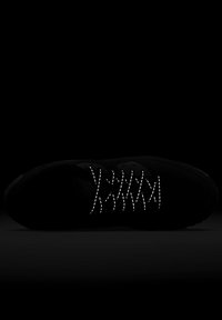 Baskets sombres avec lacets réfléchissants éclairées sur fond noir, montrant un motif croisé de lumière sur le dessus de la chaussure.
