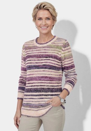 Woman with short blonde hair wearing a purple and beige striped knit sweater and beige pants, smiling with one hand in pocket.